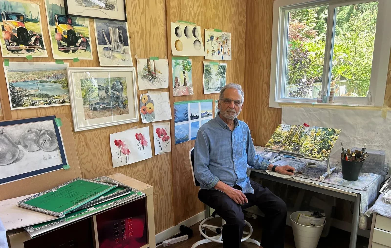 Author Mohsen Rifaat seated in his home studio surrounded by his watercolour paintings, with a paint palette and brushes on the desk beside him