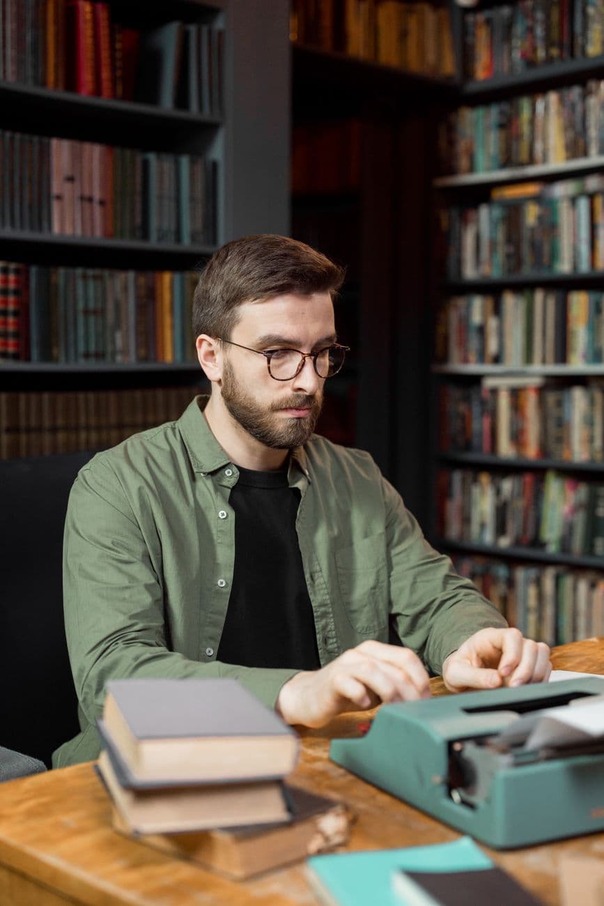 A man sits at a wooden table typing on a typewriter in a library filled with bookshelves.