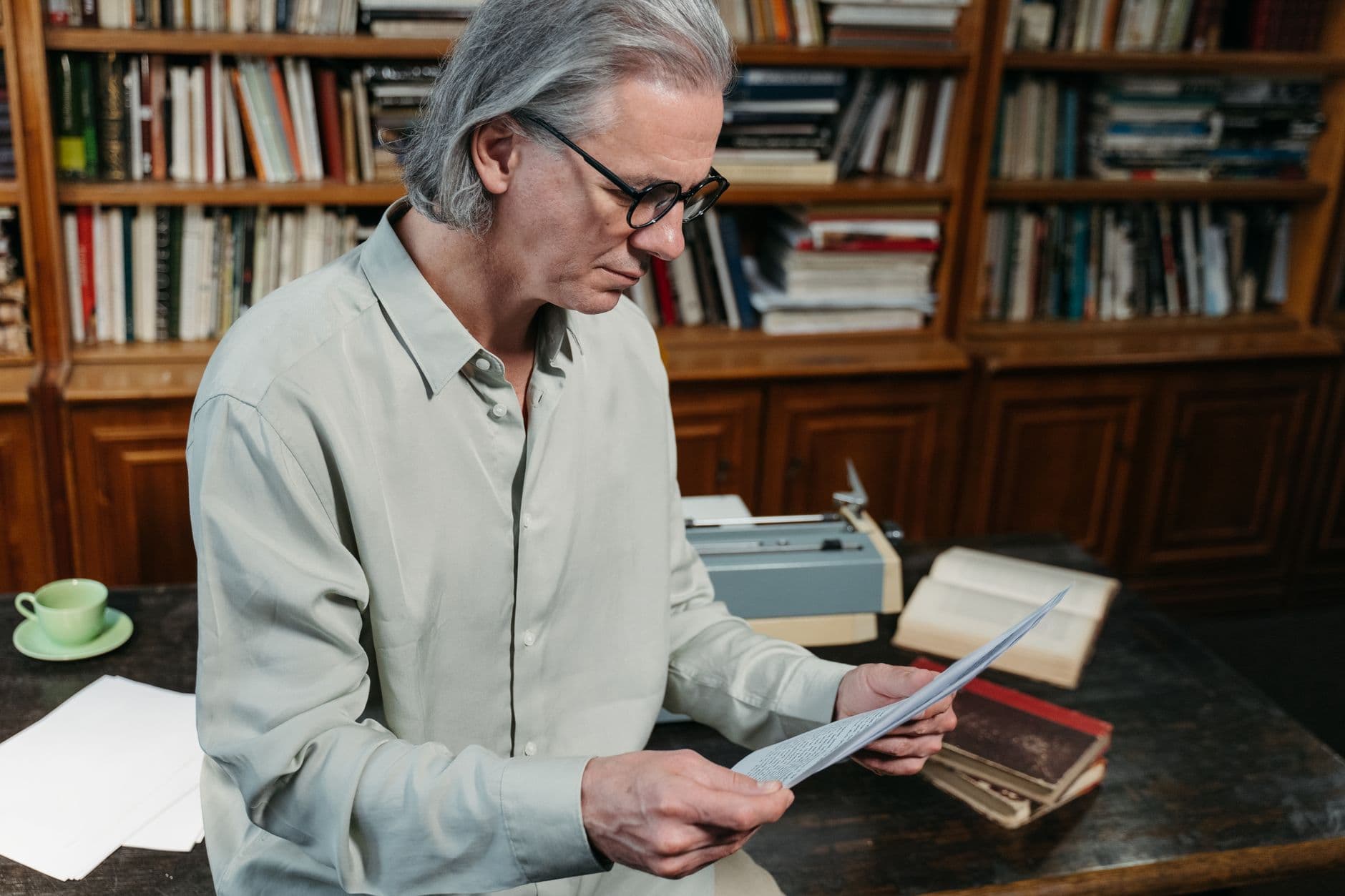 Older man in glasses reading a letter at a desk in a book-lined study, typewriter and open books beside him