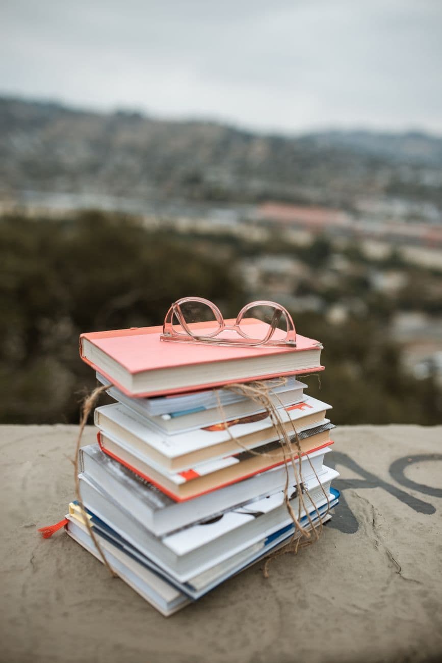 Stack of colorful books with glasses on top, set outdoors with blurred background.
